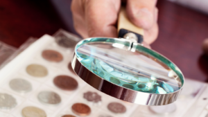 Hand holding a magnifying glass over a coin collection at Christopher Young Coins in Ithaca New York.